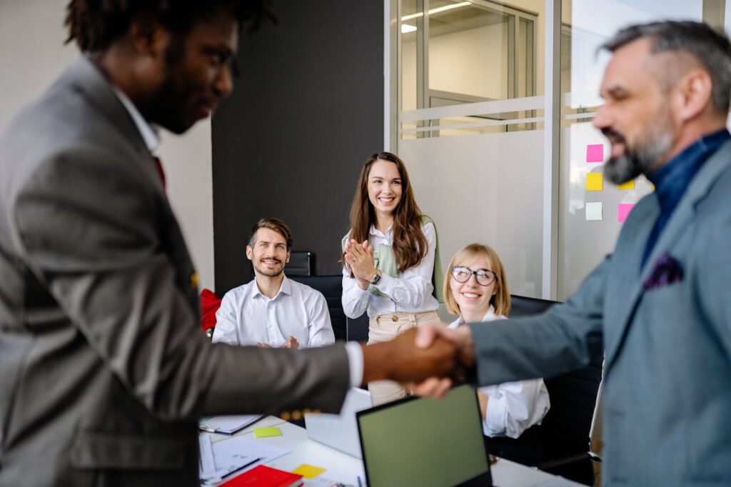 Colleagues in an office cheer as two team members finalize a successful handshake agreement.